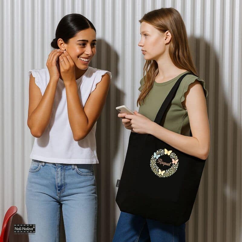 Dos chicas sonrientes conversan mientras una de ellas lleva un bolso negro personalizado con mariposas y el nombre Raquel, diseño artesanal de Noli-Nolina.